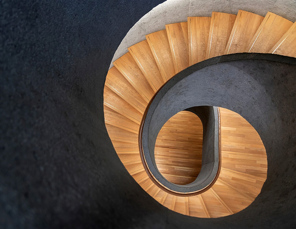 a light brown wooden spiral staircase against slate walls.